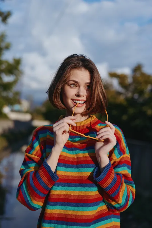 Young adult woman smiling outdoors in a colorful striped sweater, holding sunglasses by her mouth, short hair, relaxed casual outfit and playful travel lifestyle portrait.