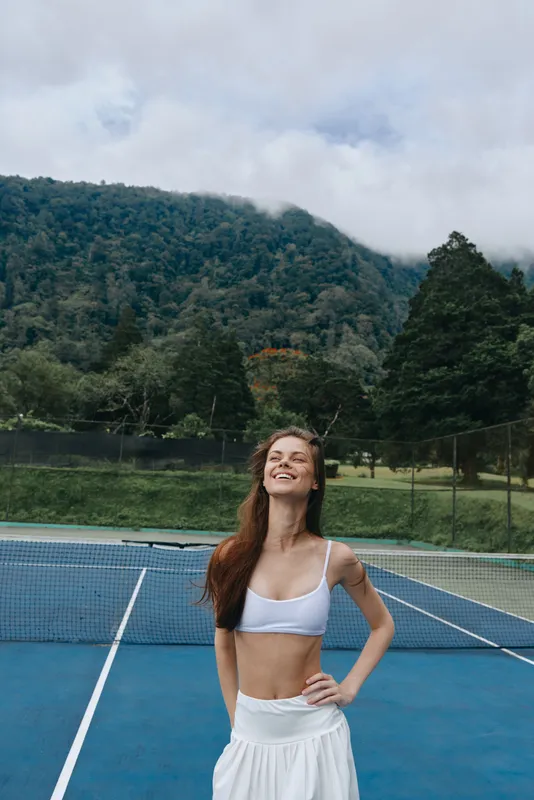 Smiling athletic woman in a sports bra and skirt on a tennis court against a greenery background, embodying healthy lifestyle and confidence