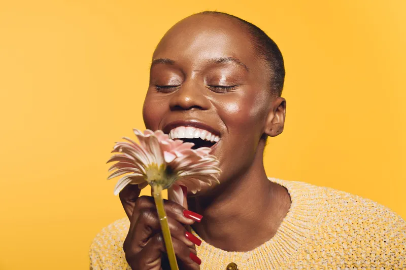 Happy African woman with dark skin smiling with closed eyes holding a pink flower near her face. Yellow background and cozy sweater create warm and joyful mood concept of beauty and nature.