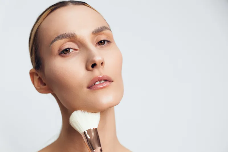 Close up portrait of woman applying makeup with brush on face, natural skin, neutral expression, clean background, soft lighting, beauty routine concept