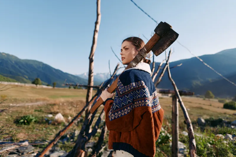 Woman carrying axe on shoulder in rural mountains landscape, wearing patterned sweater outdoors. Rustic woodcutting and homestead activity, firewood gathering in nature meadow scene.
