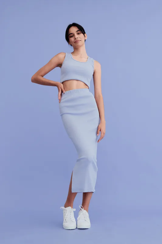 Young woman wearing stylish light blue crop top and skirt standing against solid blue background, posing confidently with casual white sneakers in modern minimalistic studio.