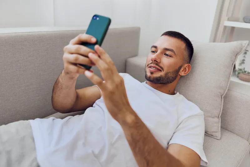 Relaxed man using smartphone on a comfortable couch, engaging with technology in a modern living space, conveying a feeling of leisure and connection.
