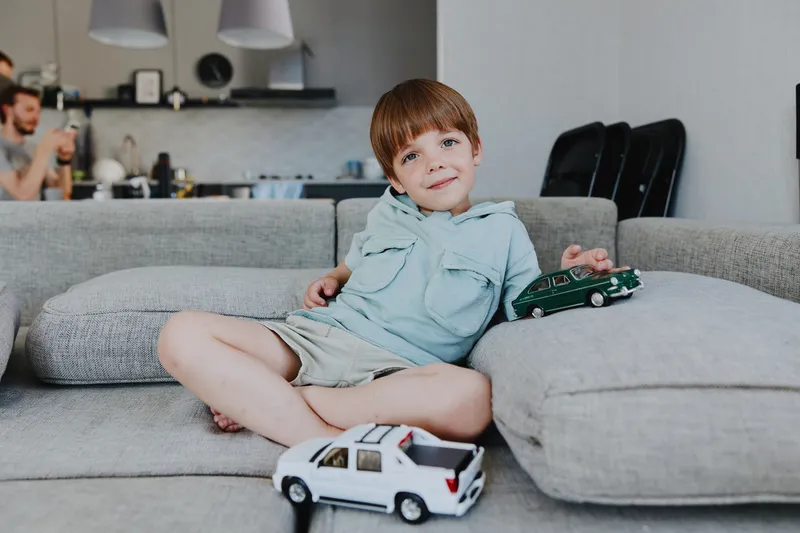 Happy boy playing with toy cars on a cozy sofa, enjoying indoor playtime. His playful expression and casual outfit create a cheerful atmosphere.