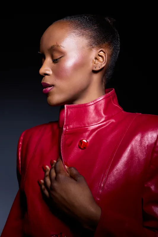 Elegant African woman with dark skin wearing a red leather jacket poses with closed eyes and hand on chest against dark background. Concept for fashion, beauty, and confidence.