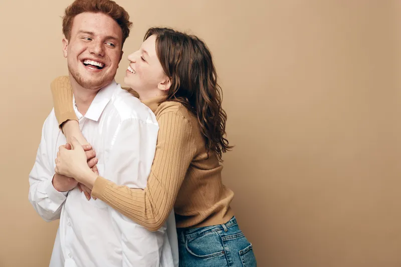 Happy young couple embraces and laughs together on beige background. Casual clothing and joyful expressions create a warm, loving atmosphere. Concept of friendship and affection.