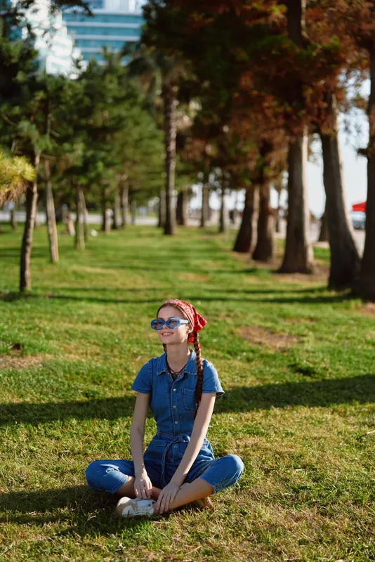 Lifestyle woman in modern boho-western denim street style sitting cross-legged on grass, wearing sunglasses and red headscarf, sunny outdoor park with trees and urban background.