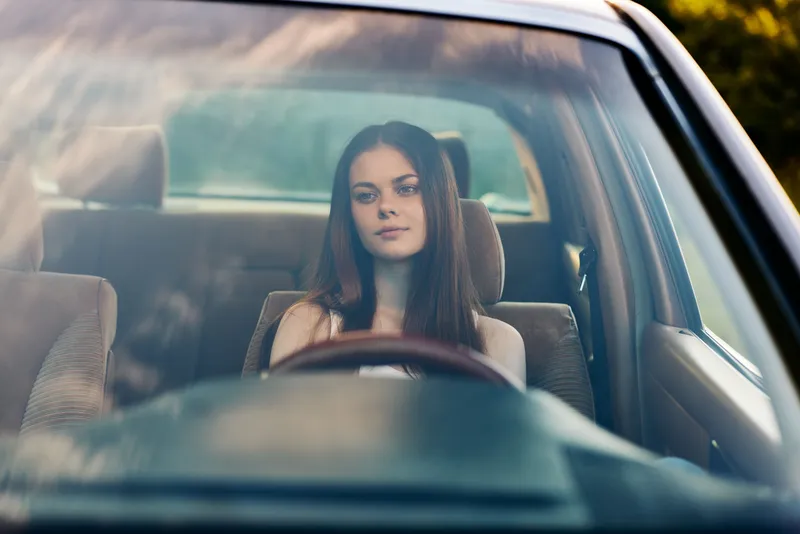 Young woman with long brown hair sitting in a car, conveying a sense of contemplation and calmness, surrounded by nature and soft sunlight.