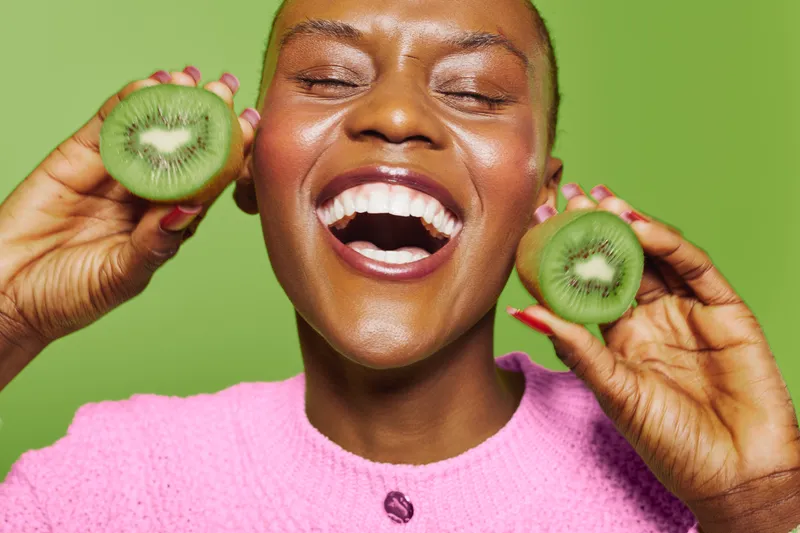 Happy dark skinned African woman laughs holding two kiwi halves near face against green background. Concept of healthy lifestyle, fresh fruit, joy, and natural beauty with vibrant colors.