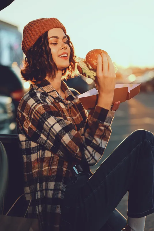 Woman eating a burger outdoors at golden hour, wearing a beanie and plaid shirt, smiling on a street bench with sunlight. Authenticity, candid lifestyle, golden hour glow, mindful living, emotional