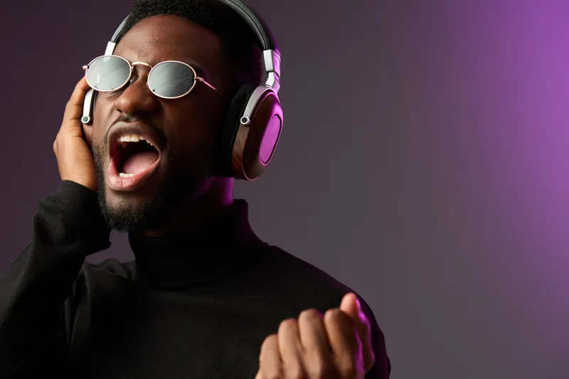 Young Black man singing passionately with headphones, wearing sunglasses and black turtleneck, colorful purple background, capturing the essence of music and emotion