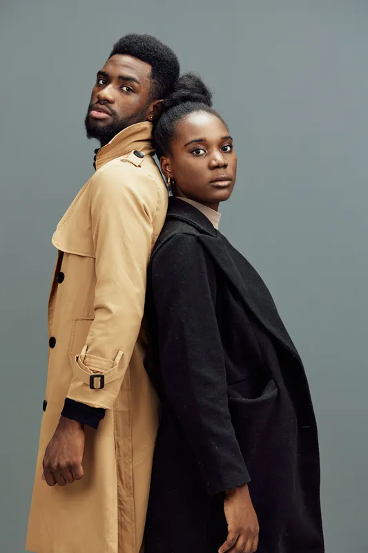 Stylish African couple standing back to back, expressing confidence and elegance in contemporary outerwear against a muted grey backdrop