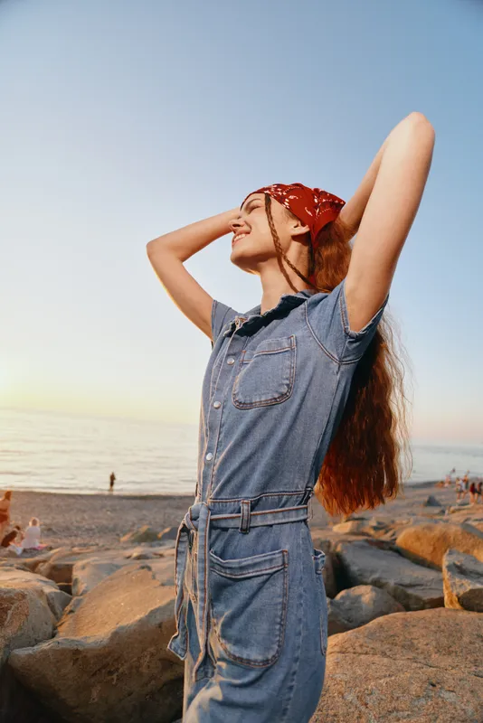 Lifestyle woman in modern boho-western denim street style enjoys sunset on rocky beach with film color tones, relaxed and carefree mood captured in natural light.
