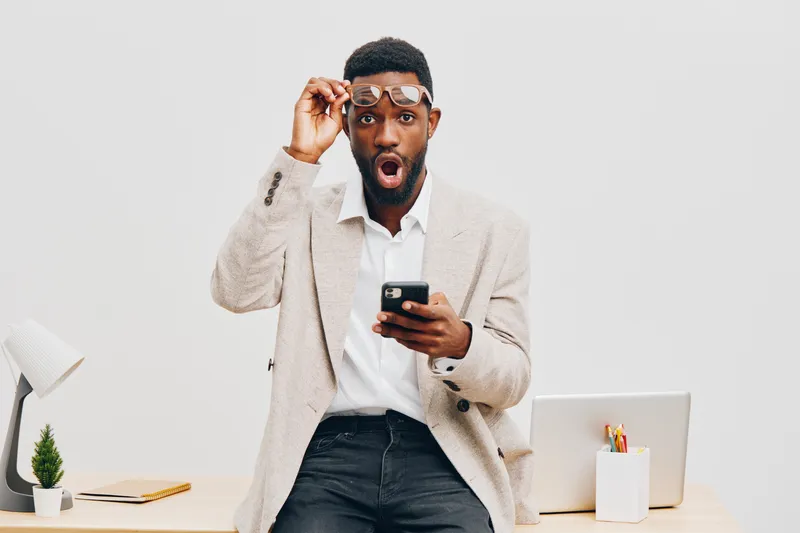 Surprised young African American man in formal outfit holding smartphone and glasses, reacting with shock, standing in modern office environment with white background, concept of surprise or shock. Pe