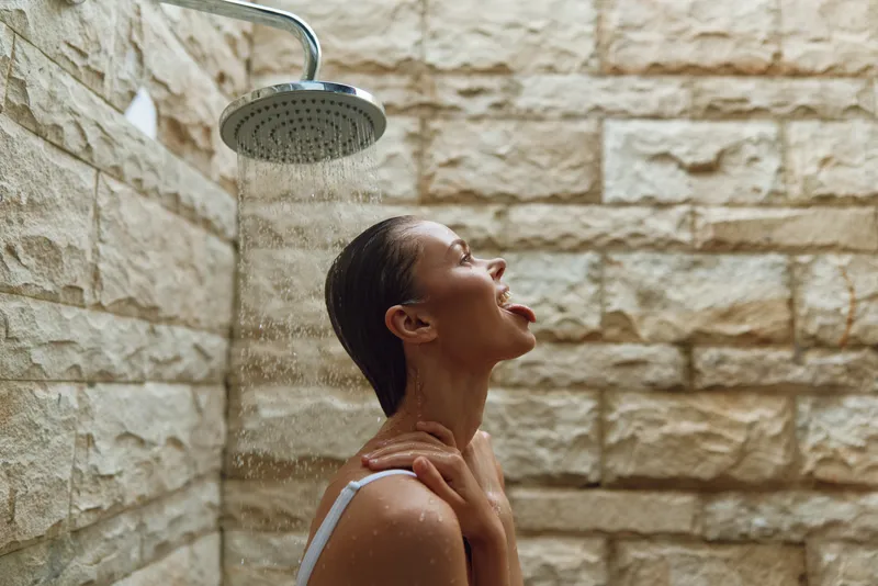 Woman enjoying refreshing shower water under rain showerhead with textured stone wall background, relaxed mood, wet dark hair, indoor bathroom setting.