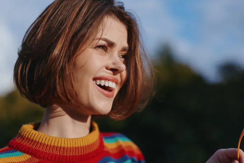 woman smiling striped sweater bob haircut outdoor natural light joyful closeup of a young adult model (mid 20s) in a colorful casual outfit; candid autumn lifestyle portrait for travel and fashion