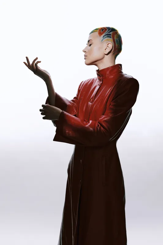Woman with colorful shaved hairstyle wearing a long red leather coat posing gracefully against a plain light background with stylish and futuristic appeal.