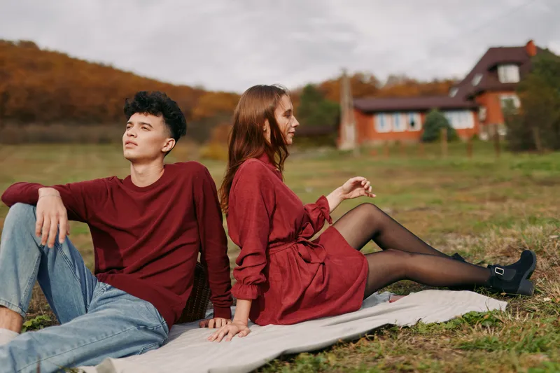A young couple sits back-to-back on a blanket in a rural field, relaxed and naturally posed, conveying a genuine connection, with authenticity and credibility in a candid outdoor portrait.