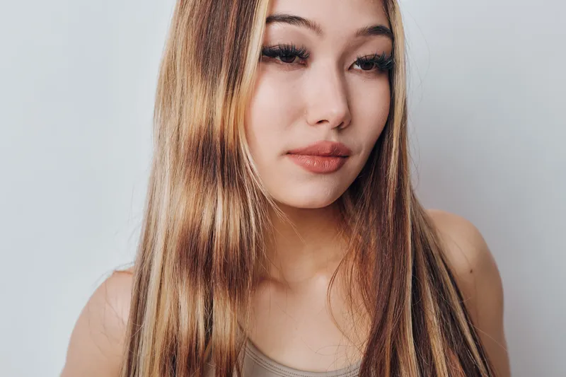 Portrait of young woman with long straight hair and natural makeup looking down with neutral expression against plain background. Closeup of thoughtful female model with smooth skin.