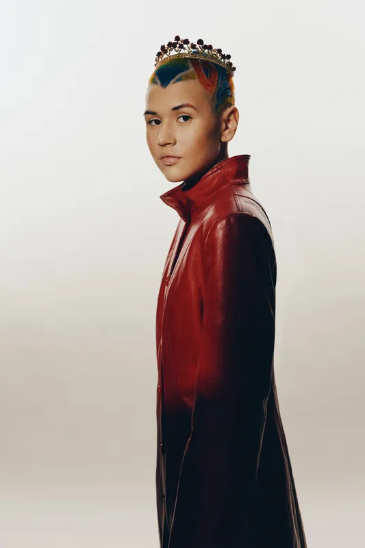 Woman with multicolored hair wearing crown and dark red leather jacket standing against light background, showcasing bold fashion and creative self-expression.