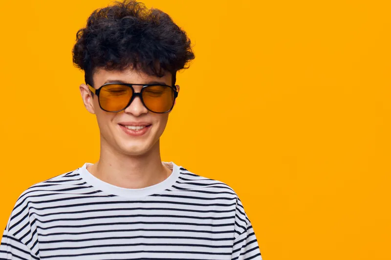 Smiling man with curly hair wearing large tinted sunglasses and a striped shirt against a vibrant orange background, showcasing a casual urban style.