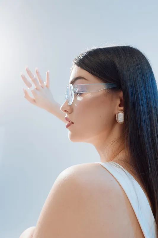 Profile of a beautiful woman with long dark hair wearing clear glasses and white top, reaching out with her hand towards bright light. Concept of beauty, fashion, and elegance.