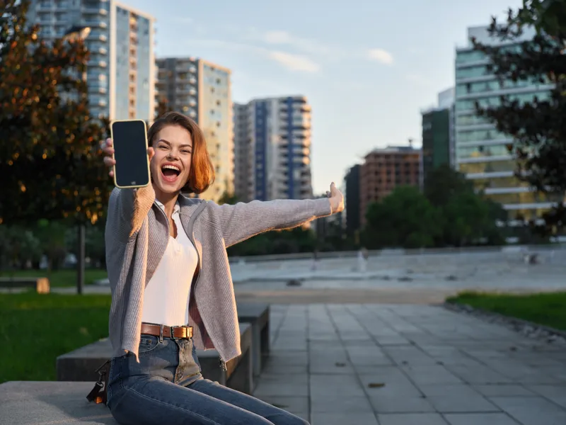 A cheerful woman takes a selfie outdoors in a modern urban park. She smiles warmly, arms outstretched, exuding confidence, energy, and a positive lifestyle in a vibrant city setting.