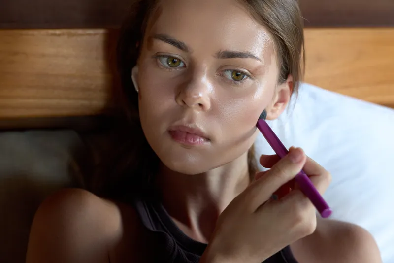 A woman applies makeup with a purple pencil in bed, capturing a calm morning beauty routine in a cozy bedroom, a closeup scene focused on selfcare, relaxation, and personal grooming.