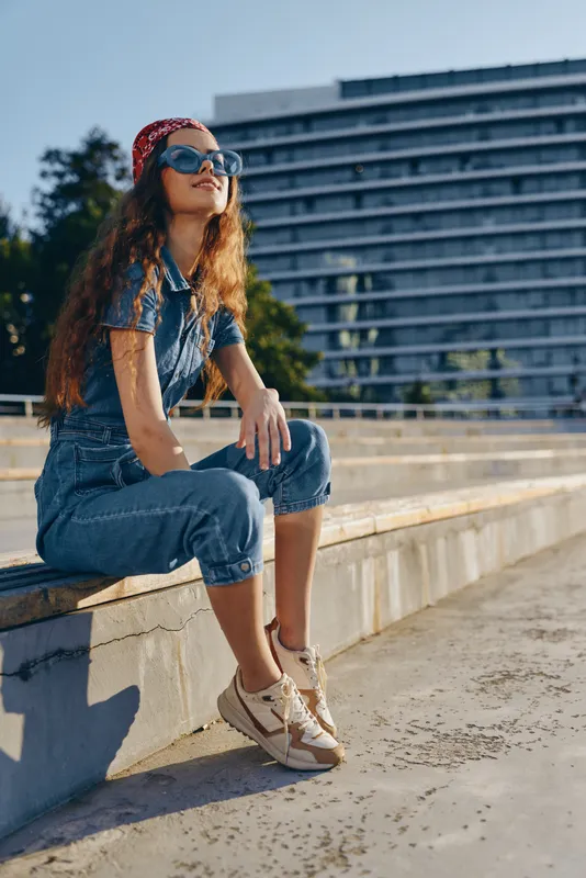 lifestyle woman in modern boho-western denim street style wearing sunglasses and sneakers, sitting outdoors with film color tones under sunlight