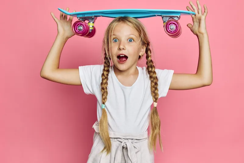 Young girl with braided hair holds a blue skateboard above her head with a surprised expression on a pink background. Fun and playful concept.