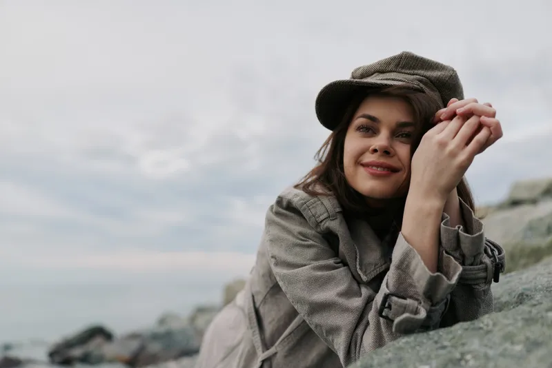 Young woman in grey coat and hat leans on rocks by the sea, smiling softly. Overcast sky and calm water create a peaceful outdoor mood at the coastline. Concept of relaxation and nature.