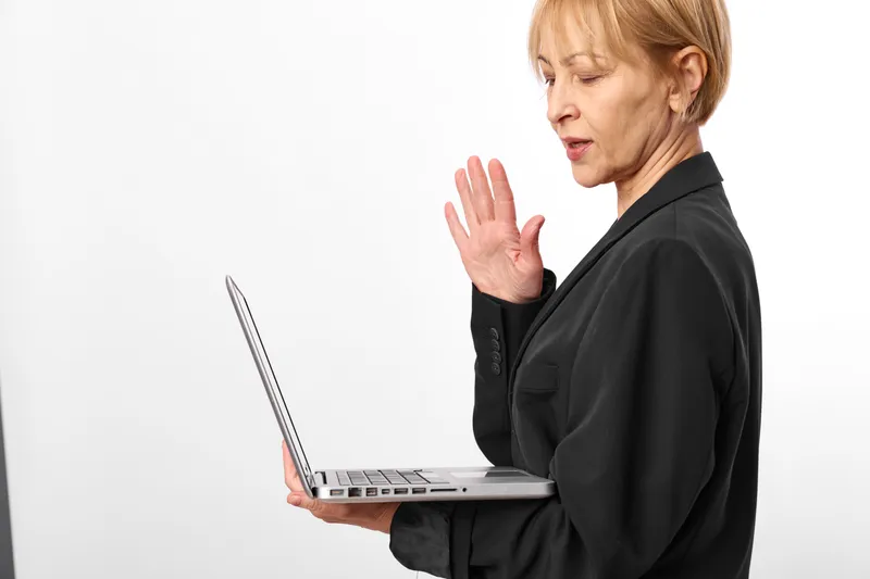 Business woman with laptop, raising hand for waving, focused expression, indoors on white background studio, using technology for communication, work mood, office setting