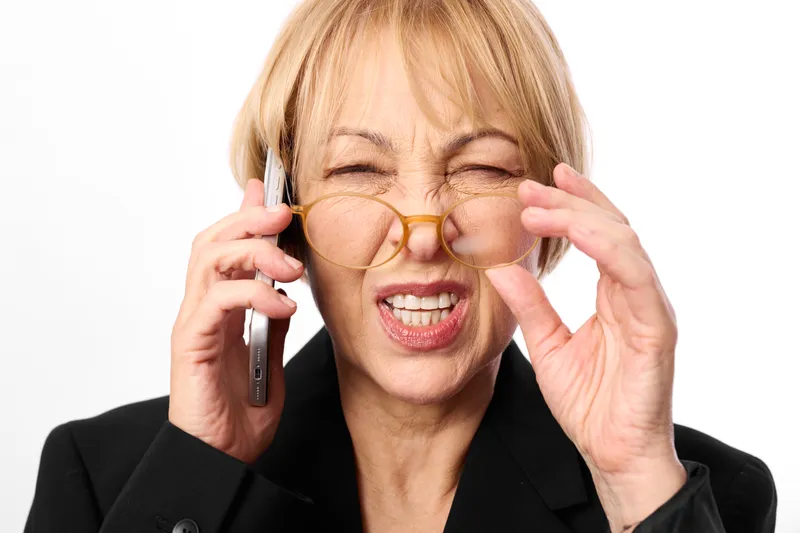 Senior woman in business suit holds phone to ear, adjusts glasses for laughter, with stress, communication and contact in bright studio background