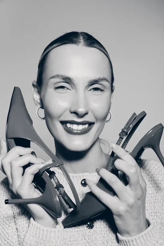 Smiling caucasian woman with light skin holding two high heel shoes close to her face. Concept of fashion, style, footwear, and happy shopper in casual sweater studio portrait.