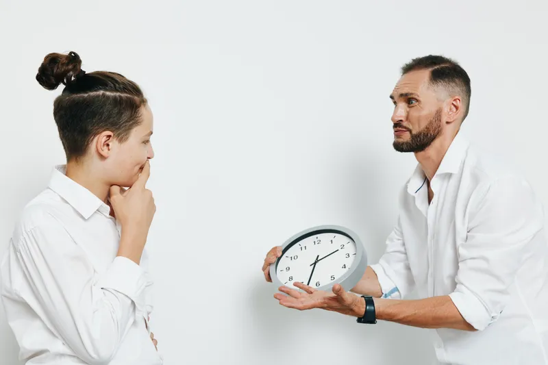 A focused male professional gestures with a clock toward a thoughtful colleague, set against an isolated colored background, conveying timing, urgency, and strategic discourse in a corporate business