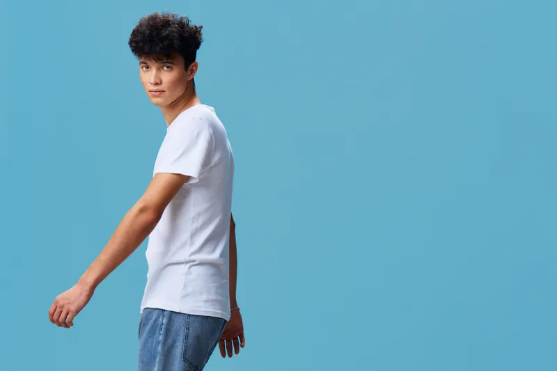 young man wearing white t-shirt and blue jeans stands confidently against blue background, casual style, curly hair, side view portrait, modern fashion look