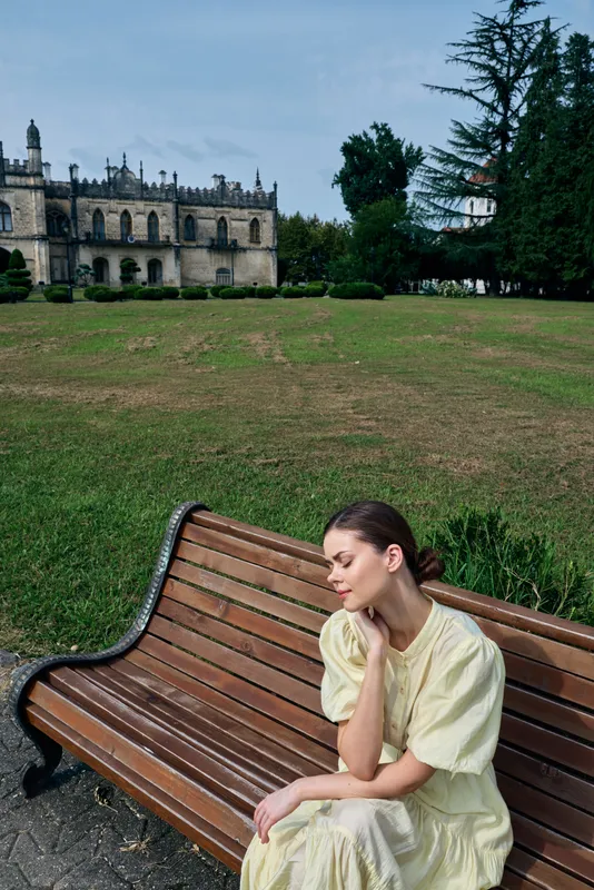 A woman sits on a wooden bench in a sunny park wearing a pale dress, lost in contemplation as a castle rises behind lush greenery, creating a serene outdoor landscape.