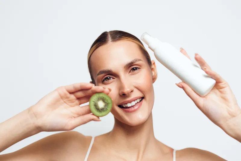 Smiling woman holding half of kiwi fruit and white cosmetic bottle, skincare concept, natural beauty, clean background, fresh healthy lifestyle, bright studio light