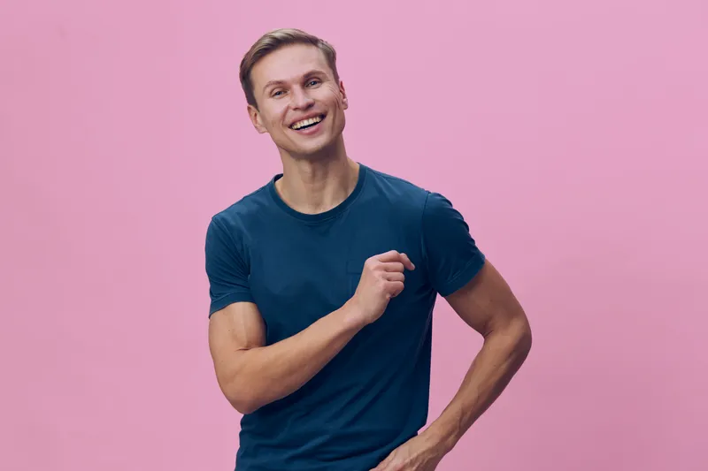 Joyful young caucasian man with short brown hair wearing casual blue t-shirt smiling happily against pink background. Male lifestyle portrait emphasizing positivity, youth, and friendliness, studio sh