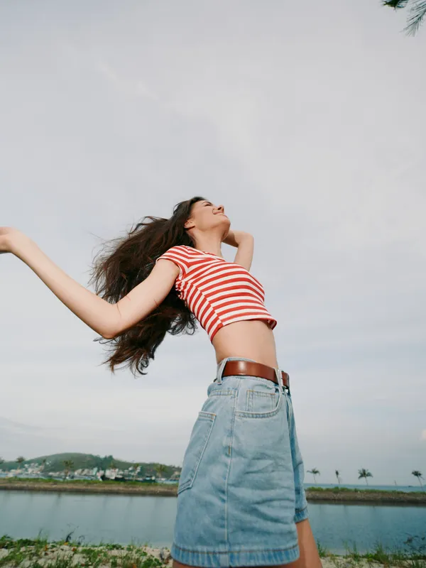 Woman wearing striped crop top and denim skirt laughs with arms outstretched as wind blows her hair, standing by a calm lake under a cloudy, expansive sky, radiating playfulness.