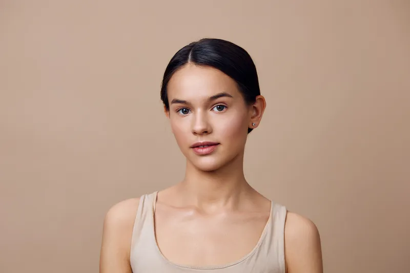 Natural portrait of a young woman with smooth skin and minimal makeup against a beige background, embodying beauty and simplicity in a fresh, seasonal look