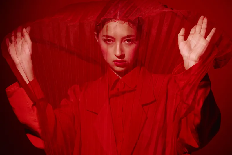 Dramatic woman in red clothing with bold makeup poses behind a sheer fabric. Artistic beauty concept with lighting creating intense mood and a fashion forward creative look.