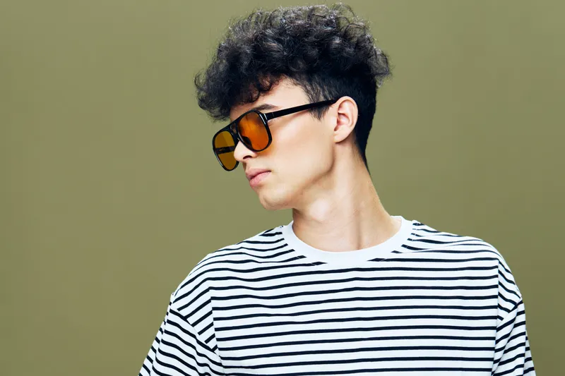 Young man with curly hair wearing striped shirt and orange sunglasses looking sideways on olive green background, casual and stylish portrait in soft lighting.