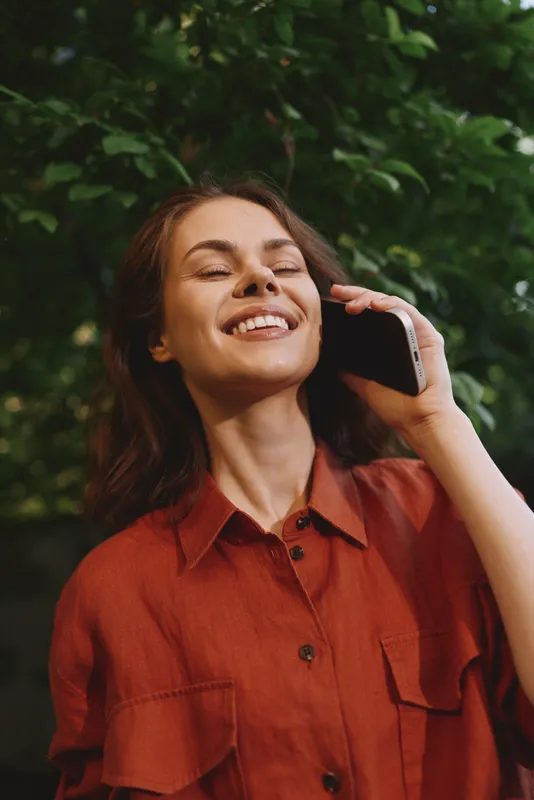 Smiling woman outdoors wearing a red shirt talks on a phone, natural light highlights greenery, a casual lifestyle moment with a warm, relaxed mood