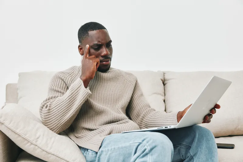 Thoughtful African man in cozy sweater working on a laptop at home, reflecting on ideas and productivity in a serene environment