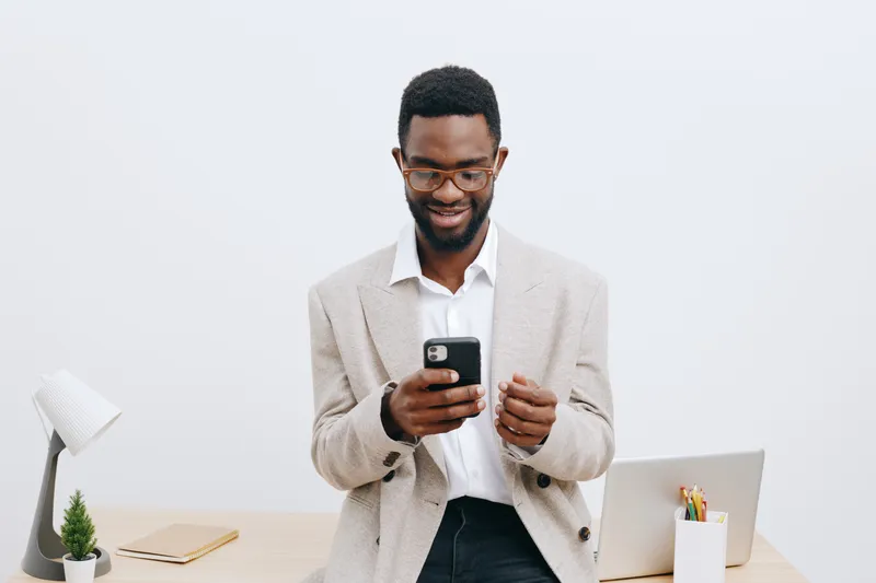 Smiling man with glasses using mobile phone while standing at work desk in modern office, casual clothing, joyful expression, contemporary workspace with laptop, stationery, and houseplant on white ba