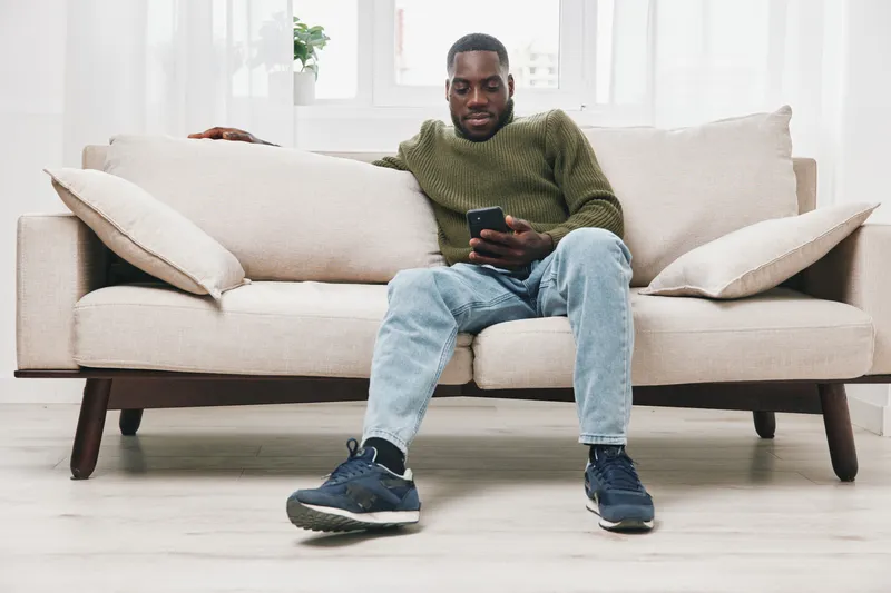 Casual Black man relaxing on a comfortable sofa, using a smartphone, wearing a cozy sweater and jeans, showcasing a calm and relaxed emotion in a bright, minimalistic room