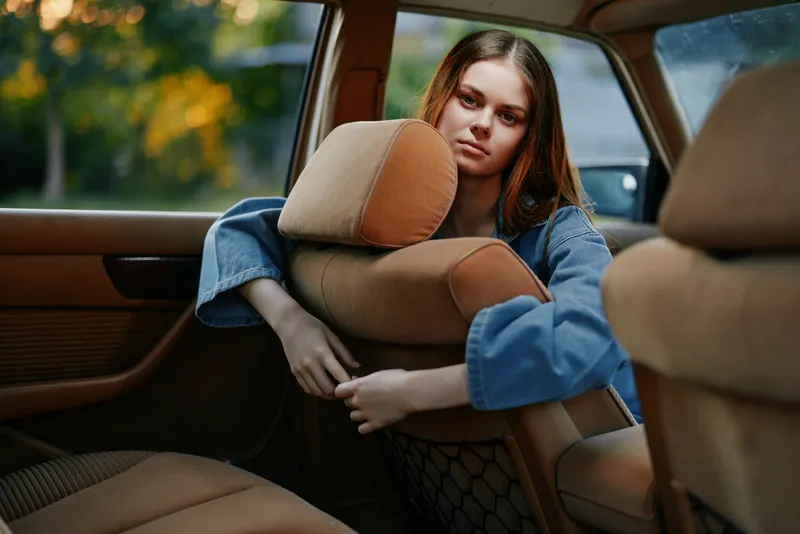 A young woman with long hair in a denim jacket looks thoughtfully out of a car window, reflecting on a sunny day, embodying feelings of solitude and contemplation.