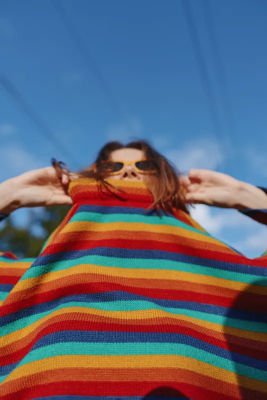 woman, striped sweater, sunglasses, blue sky, outdoors, pulling sweater — Young mid-twenties woman in a rainbow knit pulls the collar over her smile, playful low-angle portrait in sunny park with