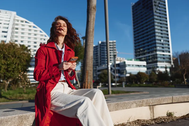 Young woman with long curly hair wearing a red coat sits on a stone bench in an urban park while smiling and holding a smartphone on a sunny day.